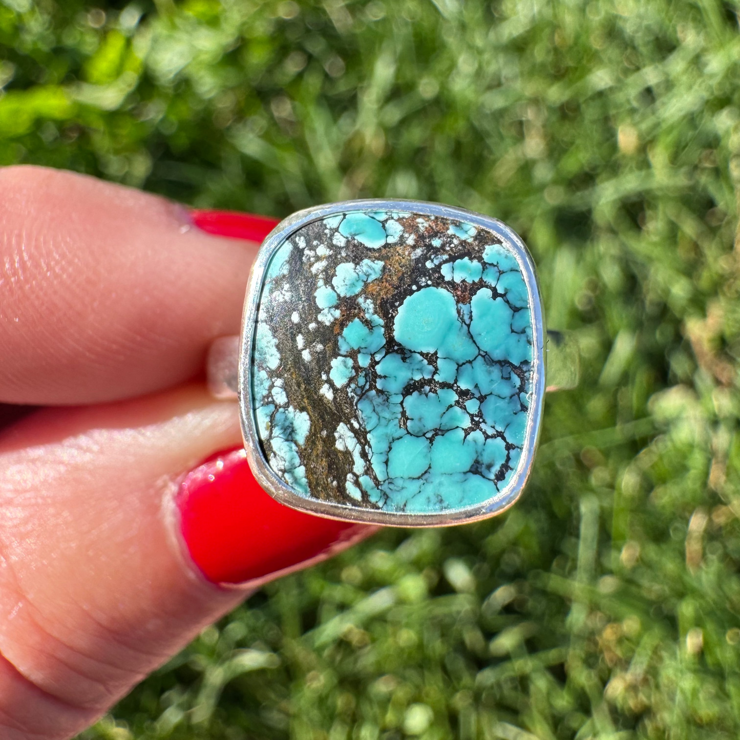 A close-up of a hand holding a square silver ring with a turquoise and brown marbled stone, set against a blurred green grass background.
