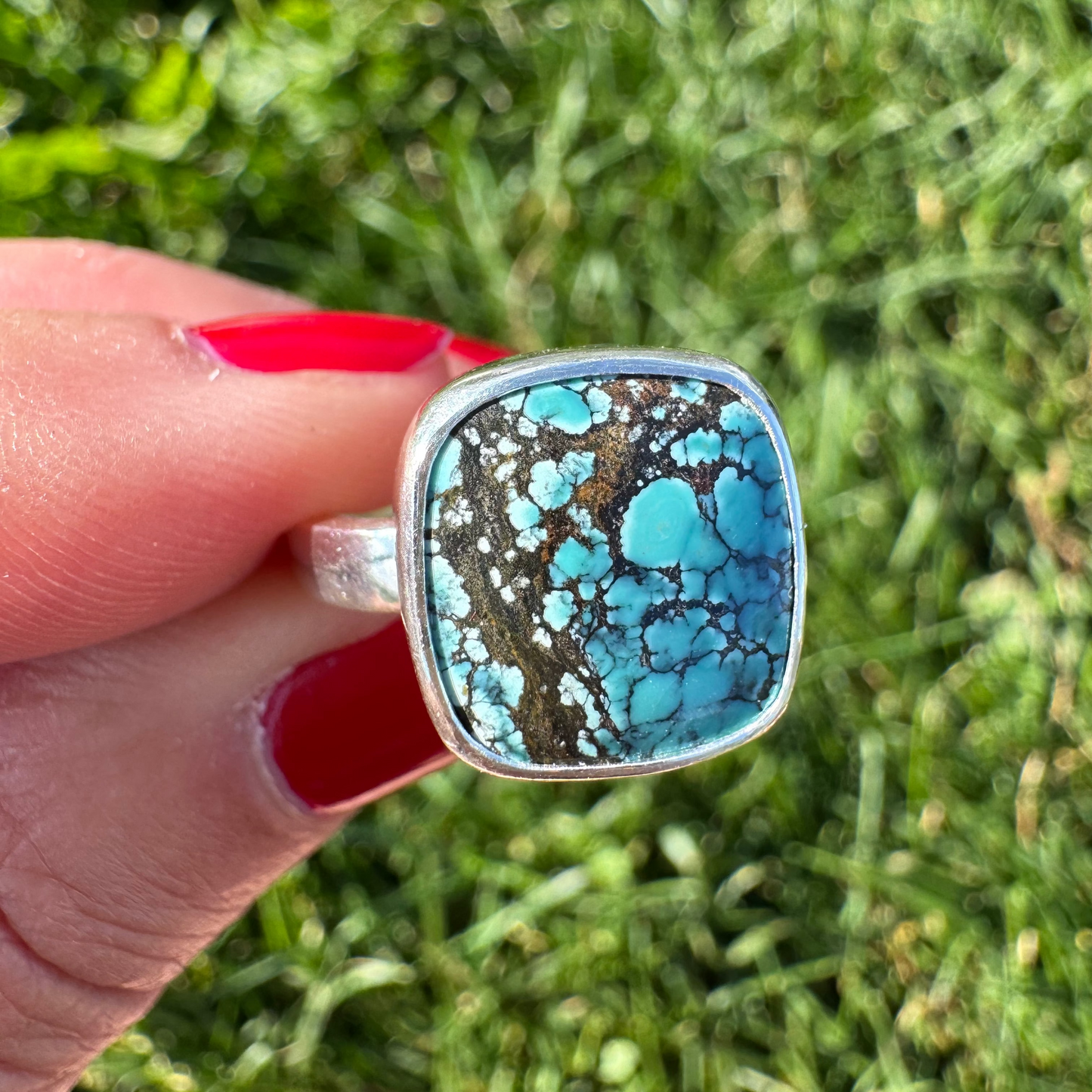A silver ring with a large square cushion-cut turquoise stone featuring a brown matrix pattern, held by a hand with red nail polish against a blurred green grass background.