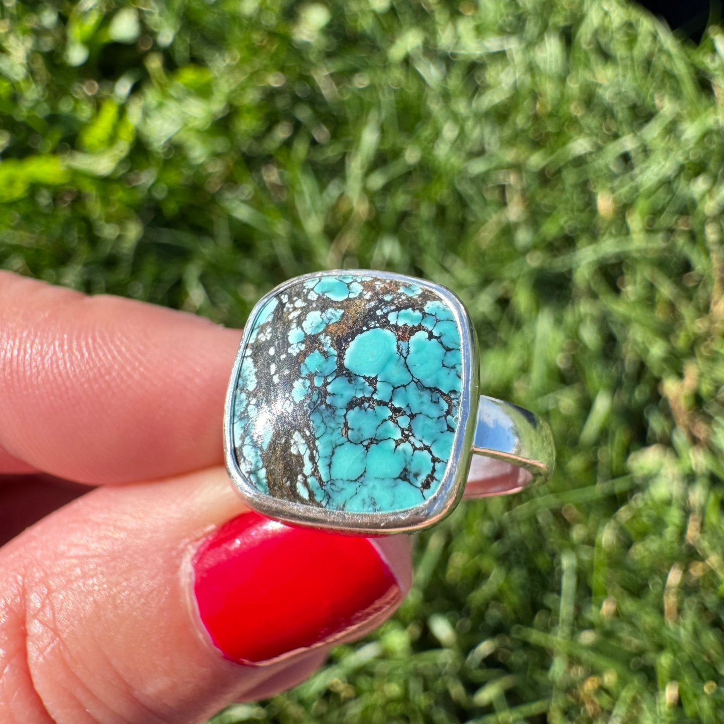 A silver ring with a large turquoise-colored stone featuring dark veining, held by a hand with red nail polish against a blurred green grass background.