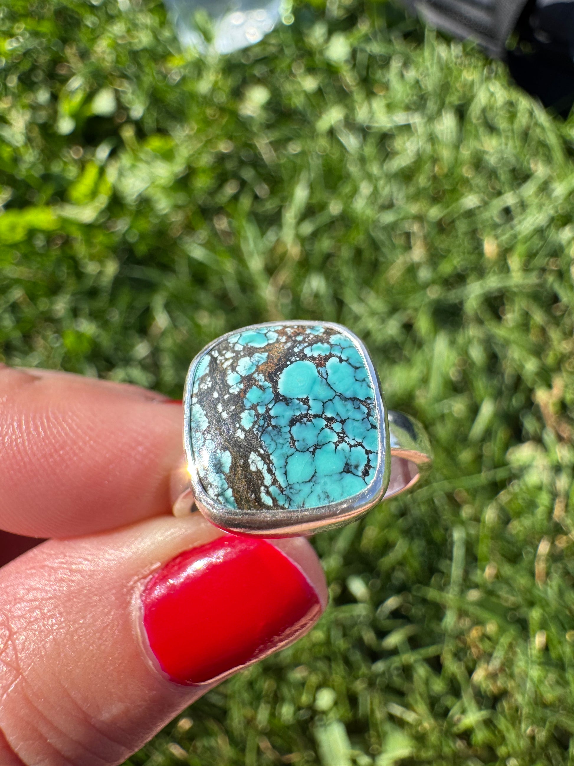 A silver ring with a large turquoise and brown marbled stone, held by a hand with red nail polish against a blurred green grass background.