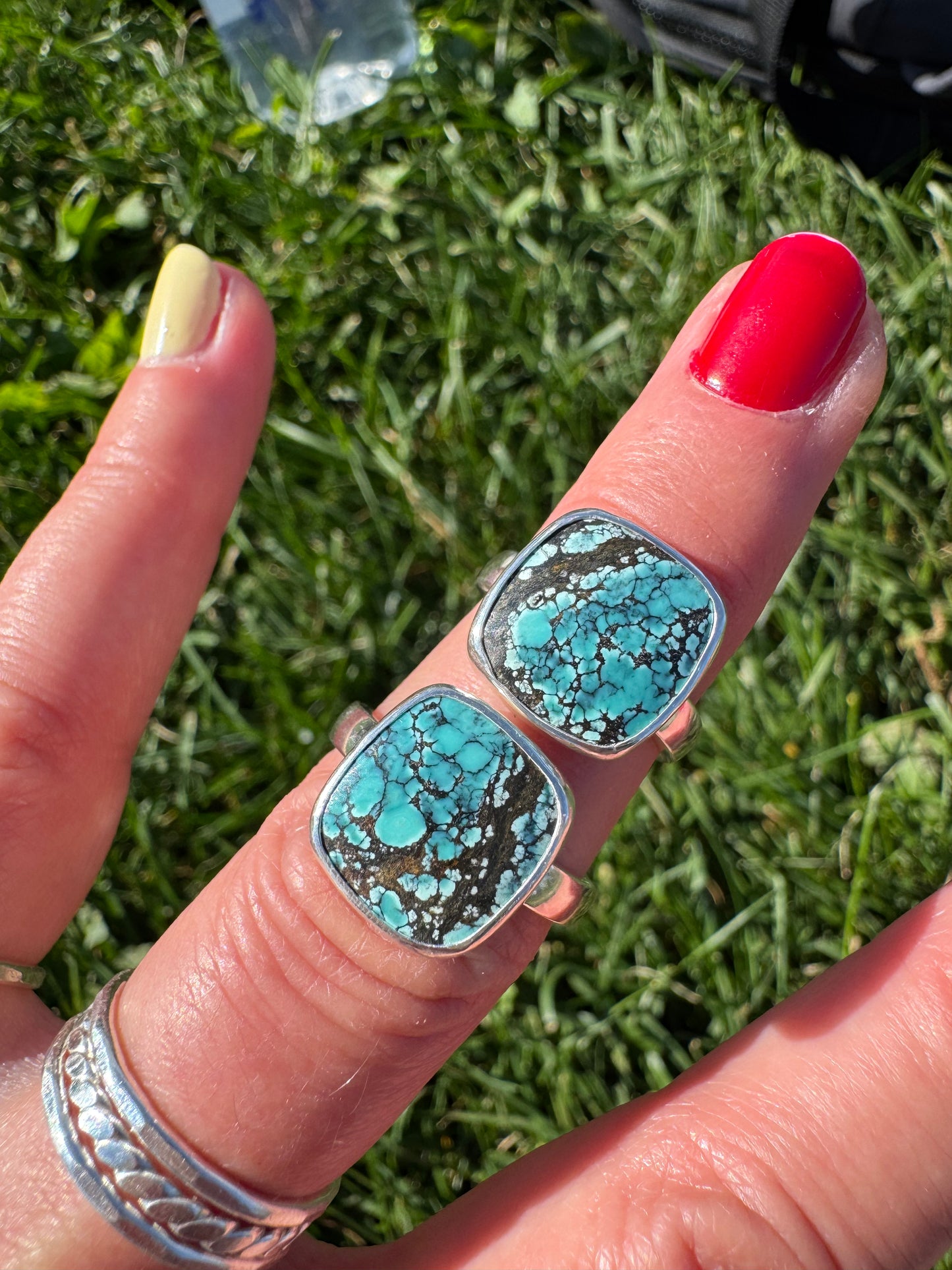 A close-up of a hand outdoors on grass wearing two large square turquoise and black marbled rings with silver-toned settings, along with a silver band on another finger.
